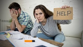 Mujer sostiene cartel que dice "Ayuda' y hombre se lleva las manos al rostro frente a papeles y calculadora.