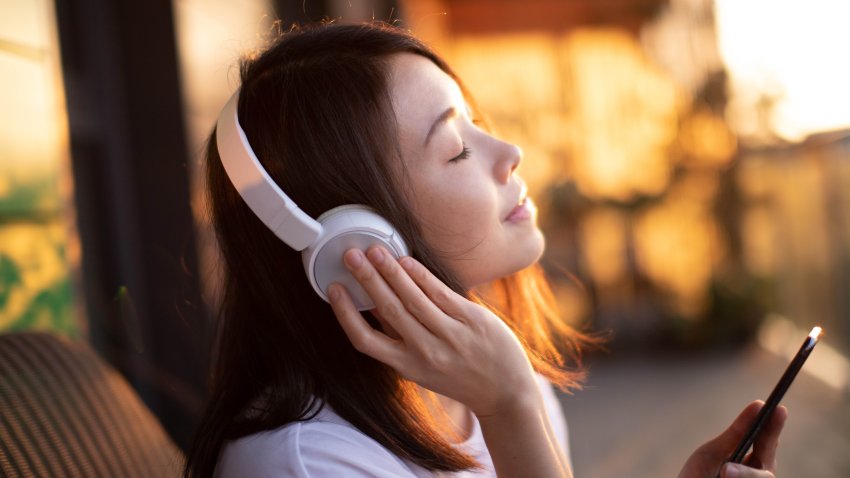 Portrait of smiling young Asian woman with eyes closed enjoying music over headphones and using smartphone, relaxing on the balcony against sunlight.