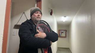 A amn in a jacket and knitted beanie stands outside and apartment holding two tennis rackets.